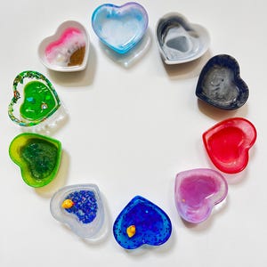 May include: A collection of heart-shaped resin dishes in various colors, including pink, blue, green, and red. Some dishes have decorative elements like glitter and small yellow ducks. The dishes are arranged in a circle on a white surface.