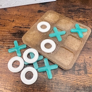 Könnte beinhalten: Ein braunes, hölzernes Tic-Tac-Toe-Spielbrett mit weißen und türkisfarbenen Spielsteinen. Das Brett liegt auf einem Holztisch. Die Spielsteine liegen verstreut um das Brett herum.