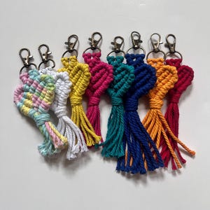 May include: A selection of handmade macrame keyrings in a variety of colours: rainbow, white, yellow, pink, teal, blue, orange, and red. Each keyring has a metal clasp for attaching to keys or bags. The keyrings are displayed on a white surface.
