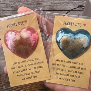 Może przedstawiać: Dwa małe, sercowate ozdoby z żywicy z wbudowanymi muszelkami i piaskiem. Każde serce jest zapakowane na kartce z tekstem "Pocket Hug" i wiadomością o trzymaniu serca blisko dla pocieszenia.