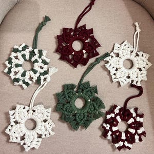 May include: Six handmade macrame snowflake ornaments in various colors: white, green, and burgundy. Each snowflake has a circular center and is attached to a hanging loop. These decorative ornaments are perfect for holiday decor.