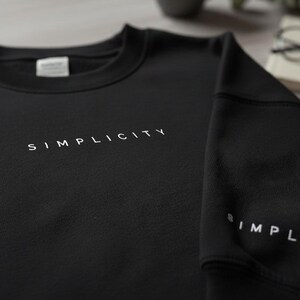 Puede incluir: Sudadera negra de cuello redondo con la palabra "SIMPLICITY" impresa en blanco en el pecho. La misma palabra también está impresa en la manga. De diseño minimalista, la sudadera está hecha de un material suave.