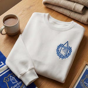 Op de afbeelding: Een witte sweatshirt met een blauw geborduurd embleem, een blauw-witte sjaal en een ingelijste print. Een kopje thee en een gevouwen beige trui liggen op een houten oppervlak. De sweatshirt heeft een ronde hals en lange mouwen.
