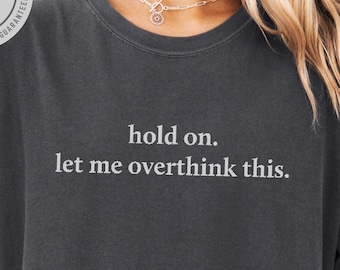 May include: Dark gray t-shirt with the text "hold on. let me overthink this." in white. The shirt has rolled-up sleeves and a relaxed fit. The color is pepper.