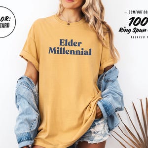 Elder Millennial graphic tee in Graphite color, made from Comfort Colors® C1717 shirt. The design features bold text in vintage-style font with 'Elder Millennial' printed in mustard yellow across the front. Available in multiple colors including Ivory, Espresso, Yam, Denim, Hemp, Blue Jean, Graphite, and Mustard. A perfect t-shirt for millennials embracing their unique experiences, including the 90s and early 2000s nostalgia.