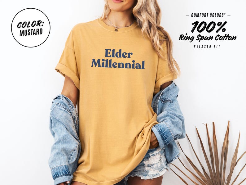 Elder Millennial graphic tee in Graphite color, made from Comfort Colors® C1717 shirt. The design features bold text in vintage-style font with 'Elder Millennial' printed in mustard yellow across the front. Available in multiple colors including Ivory, Espresso, Yam, Denim, Hemp, Blue Jean, Graphite, and Mustard. A perfect t-shirt for millennials embracing their unique experiences, including the 90s and early 2000s nostalgia.
