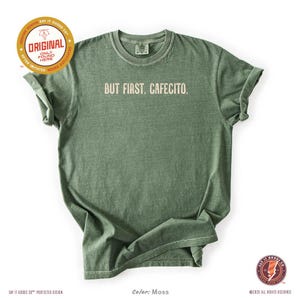 K&ouml;nnte beinhalten: Ein moosgr&uuml;nes T-Shirt mit dem wei&szlig;en Aufdruck "BUT FIRST, CAFECITO". Das T-Shirt hat einen Rundhalsausschnitt und kurze &Auml;rmel. Das Design enth&auml;lt das Logo "Say It Goods Co." und den Text "ORIGINAL ONLY FOUND HERE".