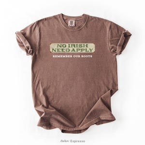 Graphic tee featuring the phrase “No Irish Need Apply” with “Remember Our Roots” printed beneath in bold white lettering. A vintage, historical design on a Comfort Colors® cotton shirt, perfect for Irish Americans, history buffs, educators, and those honoring immigrant heritage. Ideal for St. Patrick’s Day, cultural pride, or anti-discrimination messaging. Available in unisex sizing.