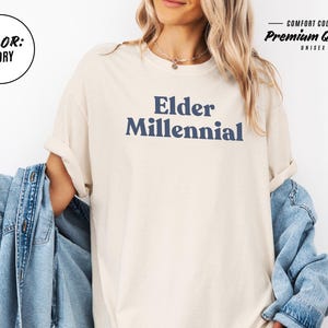 Elder Millennial graphic tee in Graphite color, made from Comfort Colors® C1717 shirt. The design features bold text in vintage-style font with 'Elder Millennial' printed in mustard yellow across the front. Available in multiple colors including Ivory, Espresso, Yam, Denim, Hemp, Blue Jean, Graphite, and Mustard. A perfect t-shirt for millennials embracing their unique experiences, including the 90s and early 2000s nostalgia.