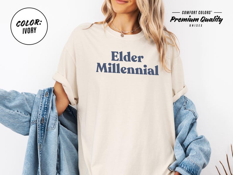 Elder Millennial graphic tee in Graphite color, made from Comfort Colors® C1717 shirt. The design features bold text in vintage-style font with 'Elder Millennial' printed in mustard yellow across the front. Available in multiple colors including Ivory, Espresso, Yam, Denim, Hemp, Blue Jean, Graphite, and Mustard. A perfect t-shirt for millennials embracing their unique experiences, including the 90s and early 2000s nostalgia.