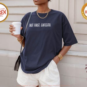 K&ouml;nnte beinhalten: Marineblaues Unisex-T-Shirt mit dem wei&szlig;en Text "BUT FIRST. CAFECITO.". Das Shirt besteht aus 100 % Baumwolle und ist in den Gr&ouml;&szlig;en S-4XL erh&auml;ltlich. Das Model tr&auml;gt wei&szlig;e Shorts und h&auml;lt einen wei&szlig;en Kaffeebecher.