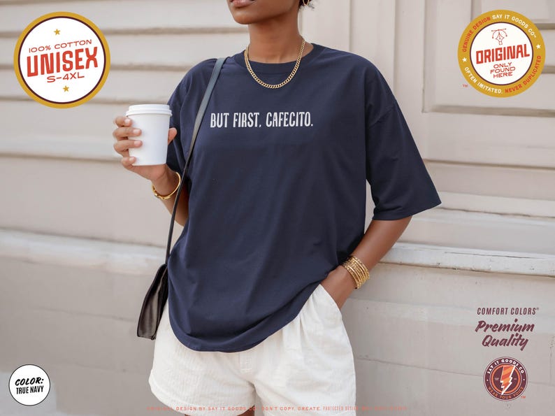 K&ouml;nnte beinhalten: Marineblaues Unisex-T-Shirt mit dem wei&szlig;en Text "BUT FIRST. CAFECITO.". Das Shirt besteht aus 100 % Baumwolle und ist in den Gr&ouml;&szlig;en S-4XL erh&auml;ltlich. Das Model tr&auml;gt wei&szlig;e Shorts und h&auml;lt einen wei&szlig;en Kaffeebecher.