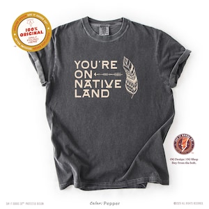 Puede incluir: Una camiseta gris oscuro con el texto "YOU'RE ON NATIVE LAND" en color beige claro, junto con un diseño de flecha y pluma. La camiseta tiene un aspecto vintage y desgastado e incluye el texto "100% ORIGINAL" en una pegatina.