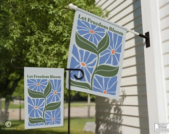 May include: Two garden flags with the text "Let Freedom Bloom". The flags feature a blue floral design with green leaves and orange centers. One flag is mounted on a pole, the other on a black metal stand.