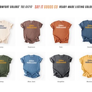 Elder Millennial graphic tee in Graphite color, made from Comfort Colors® C1717 shirt. The design features bold text in vintage-style font with 'Elder Millennial' printed in mustard yellow across the front. Available in multiple colors including Ivory, Espresso, Yam, Denim, Hemp, Blue Jean, Graphite, and Mustard. A perfect t-shirt for millennials embracing their unique experiences, including the 90s and early 2000s nostalgia.