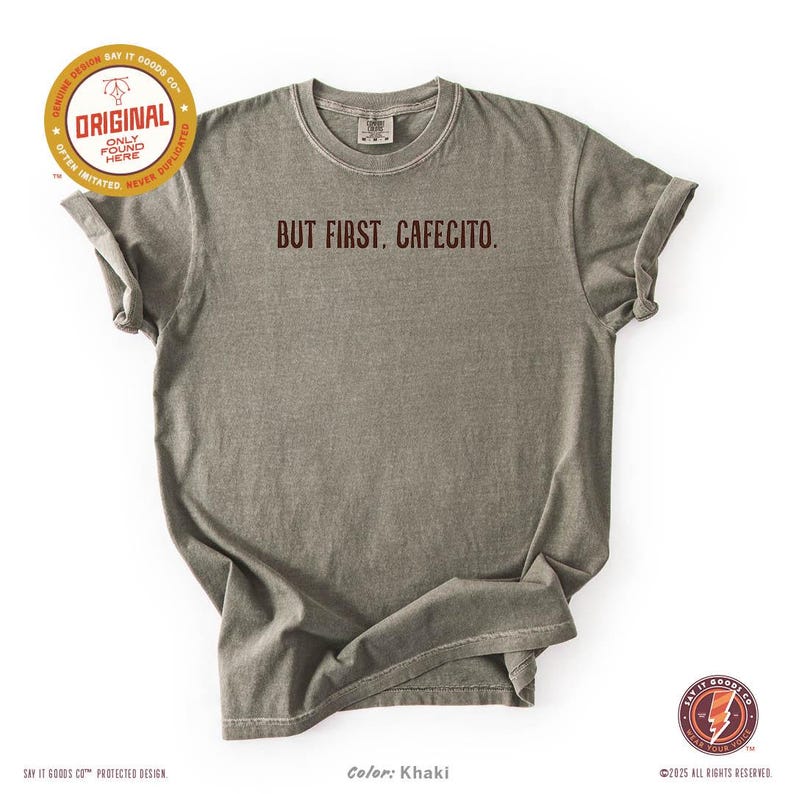 K&ouml;nnte beinhalten: Khakifarbenes T-Shirt mit dem Aufdruck "BUT FIRST, CAFECITO." in Dunkelbraun. Das Shirt hat hochgekrempelte &Auml;rmel und einen Rundhalsausschnitt. Das Design enth&auml;lt den Text "ORIGINAL" und "SAY IT GOODS CO." auf dem Shirt.