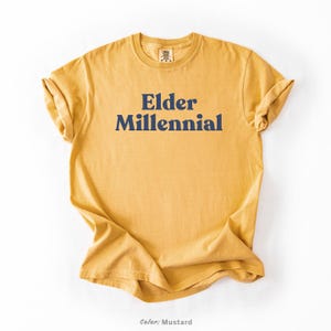 Elder Millennial graphic tee in Graphite color, made from Comfort Colors® C1717 shirt. The design features bold text in vintage-style font with 'Elder Millennial' printed in mustard yellow across the front. Available in multiple colors including Ivory, Espresso, Yam, Denim, Hemp, Blue Jean, Graphite, and Mustard. A perfect t-shirt for millennials embracing their unique experiences, including the 90s and early 2000s nostalgia.