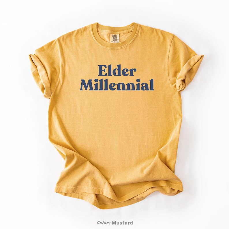 Elder Millennial graphic tee in Graphite color, made from Comfort Colors® C1717 shirt. The design features bold text in vintage-style font with 'Elder Millennial' printed in mustard yellow across the front. Available in multiple colors including Ivory, Espresso, Yam, Denim, Hemp, Blue Jean, Graphite, and Mustard. A perfect t-shirt for millennials embracing their unique experiences, including the 90s and early 2000s nostalgia.