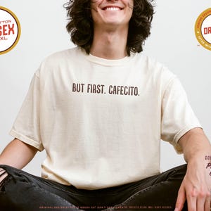 K&ouml;nnte beinhalten: Beigefarbenes Unisex-T-Shirt mit dem braunen Schriftzug "BUT FIRST, CAFECITO.". Das Shirt besteht aus 100 % Baumwolle und ist in den Gr&ouml;&szlig;en S-4XL erh&auml;ltlich. Das Bild enth&auml;lt auch Design-Details und Branding-Elemente.