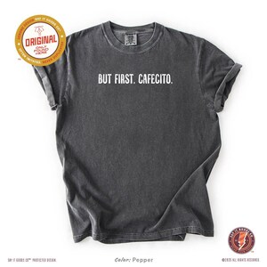 K&ouml;nnte beinhalten: Ein dunkelgraues T-Shirt mit dem wei&szlig;en Text "BUT FIRST, CAFECITO". Das Shirt hat einen Rundhalsausschnitt und kurze &Auml;rmel. Die Farbe ist Pfeffer.