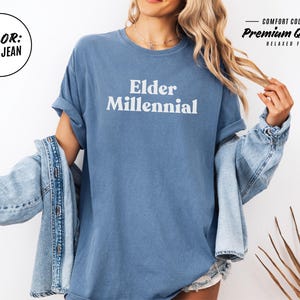 Elder Millennial graphic tee in Graphite color, made from Comfort Colors® C1717 shirt. The design features bold text in vintage-style font with 'Elder Millennial' printed in mustard yellow across the front. Available in multiple colors including Ivory, Espresso, Yam, Denim, Hemp, Blue Jean, Graphite, and Mustard. A perfect t-shirt for millennials embracing their unique experiences, including the 90s and early 2000s nostalgia.