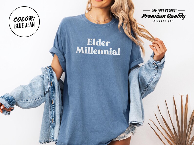 Elder Millennial graphic tee in Graphite color, made from Comfort Colors® C1717 shirt. The design features bold text in vintage-style font with 'Elder Millennial' printed in mustard yellow across the front. Available in multiple colors including Ivory, Espresso, Yam, Denim, Hemp, Blue Jean, Graphite, and Mustard. A perfect t-shirt for millennials embracing their unique experiences, including the 90s and early 2000s nostalgia.