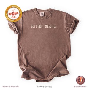Minimalist “But First. Cafecito.” graphic tee in espresso brown featuring cream typography. Latina coffee lover shirt celebrating Hispanic culture and morning cafecito ritual. Modern unisex coffee shirt perfect for espresso lovers, brunch outfits, and gift ideas for moms, sisters, and café fans.