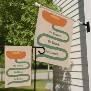 May include: Two garden flags with a floral design and the words "Be Brave. Be Kind. Be Loud." The flags feature an orange flower and a green stem on a beige background. One flag is mounted on a black stand.