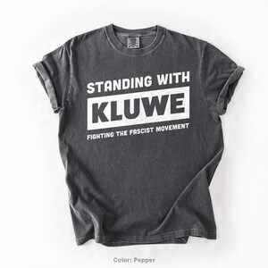 May include: A dark gray t-shirt with the text "Standing with Kluwe Fighting the Fascist Movement" printed in white.