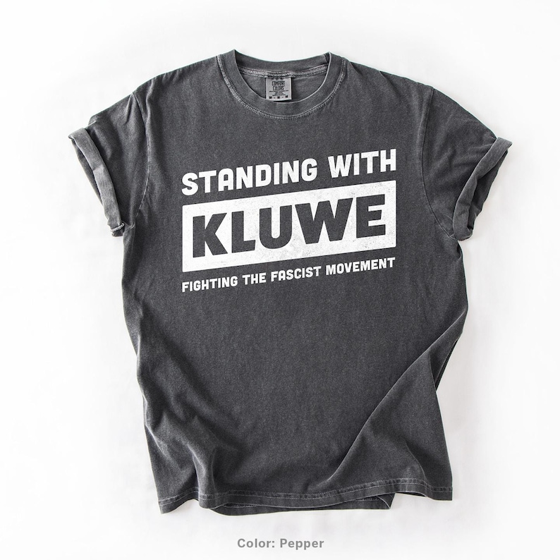 May include: A dark gray t-shirt with the text "Standing with Kluwe Fighting the Fascist Movement" printed in white.
