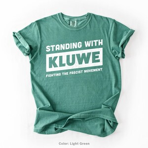 May include: A light green t-shirt with the text "Standing with Kluwe Fighting the Fascist Movement" printed in white.