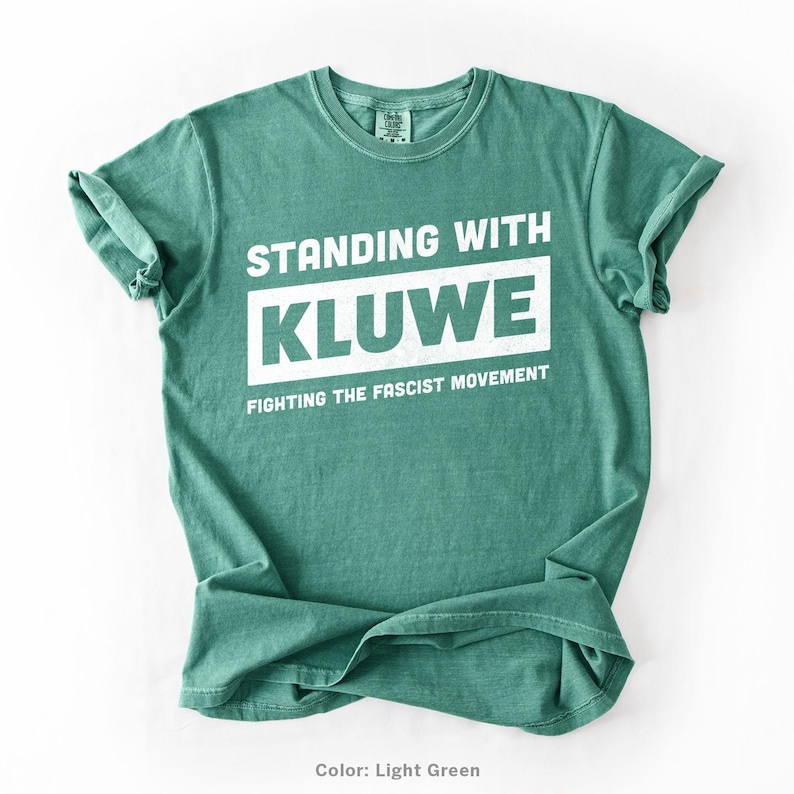 May include: A light green t-shirt with the text "Standing with Kluwe Fighting the Fascist Movement" printed in white.