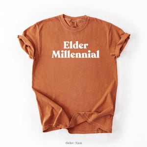 Elder Millennial graphic tee in Graphite color, made from Comfort Colors® C1717 shirt. The design features bold text in vintage-style font with 'Elder Millennial' printed in mustard yellow across the front. Available in multiple colors including Ivory, Espresso, Yam, Denim, Hemp, Blue Jean, Graphite, and Mustard. A perfect t-shirt for millennials embracing their unique experiences, including the 90s and early 2000s nostalgia.