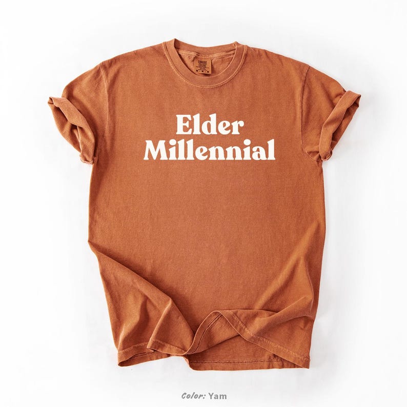Elder Millennial graphic tee in Graphite color, made from Comfort Colors® C1717 shirt. The design features bold text in vintage-style font with 'Elder Millennial' printed in mustard yellow across the front. Available in multiple colors including Ivory, Espresso, Yam, Denim, Hemp, Blue Jean, Graphite, and Mustard. A perfect t-shirt for millennials embracing their unique experiences, including the 90s and early 2000s nostalgia.