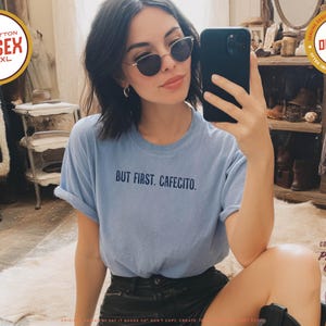 K&ouml;nnte beinhalten: Ein hellblaues Unisex-T-Shirt mit dem Text "BUT FIRST. CAFECITO." wird gezeigt. Das Shirt besteht aus 100 % Baumwolle und ist in den Gr&ouml;&szlig;en S-4XL erh&auml;ltlich. Das Shirt hat die Farbe Blue Jean.