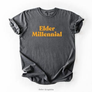 Elder Millennial graphic tee in Graphite color, made from Comfort Colors® C1717 shirt. The design features bold text in vintage-style font with 'Elder Millennial' printed in mustard yellow across the front. Available in multiple colors including Ivory, Espresso, Yam, Denim, Hemp, Blue Jean, Graphite, and Mustard. A perfect t-shirt for millennials embracing their unique experiences, including the 90s and early 2000s nostalgia.
