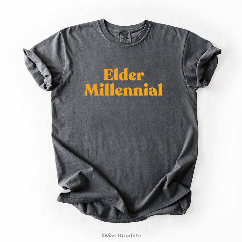 Elder Millennial graphic tee in Graphite color, made from Comfort Colors® C1717 shirt. The design features bold text in vintage-style font with 'Elder Millennial' printed in mustard yellow across the front. Available in multiple colors including Ivory, Espresso, Yam, Denim, Hemp, Blue Jean, Graphite, and Mustard. A perfect t-shirt for millennials embracing their unique experiences, including the 90s and early 2000s nostalgia.