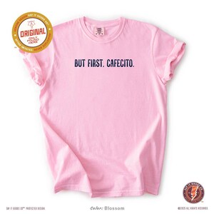 K&ouml;nnte beinhalten: Hellrosa T-Shirt mit dem marineblauen Aufdruck "BUT FIRST, CAFECITO.". Das Shirt hat kurze &Auml;rmel und einen Rundhalsausschnitt. Die Farbe wird als "Blossom" bezeichnet.