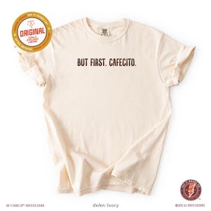 K&ouml;nnte beinhalten: Elfenbeinfarbenes T-Shirt mit dem Schriftzug "BUT FIRST, CAFECITO" in braunen Buchstaben. Das kurz&auml;rmelige T-Shirt hat einen Rundhalsausschnitt. Das Design enth&auml;lt den Text "ORIGINAL" und "SAY IT GOODS CO." auf dem Shirt.
