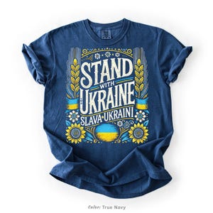 Puede incluir: Una camiseta azul marino con un diseño gráfico que presenta el texto "Stand with Ukraine" en un estilo vintage con girasoles y la bandera ucraniana. El texto "Slava Ukraini" también está incluido en el diseño.