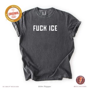 May include: A dark gray t-shirt with the text "FUCK ICE" in white block letters. The shirt has a vintage, worn look. The image includes the text "ORIGINAL" and "SAY IT GOODS CO" in a circular design.