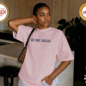 K&ouml;nnte beinhalten: Hellrosa Unisex-T-Shirt mit dem marineblauen Schriftzug "BUT FIRST. CAFECITO.". Das Shirt besteht aus 100 % Baumwolle und ist in den Gr&ouml;&szlig;en S-4XL erh&auml;ltlich. Es ist ein Premium-Qualit&auml;tsprodukt von Comfort Colors.