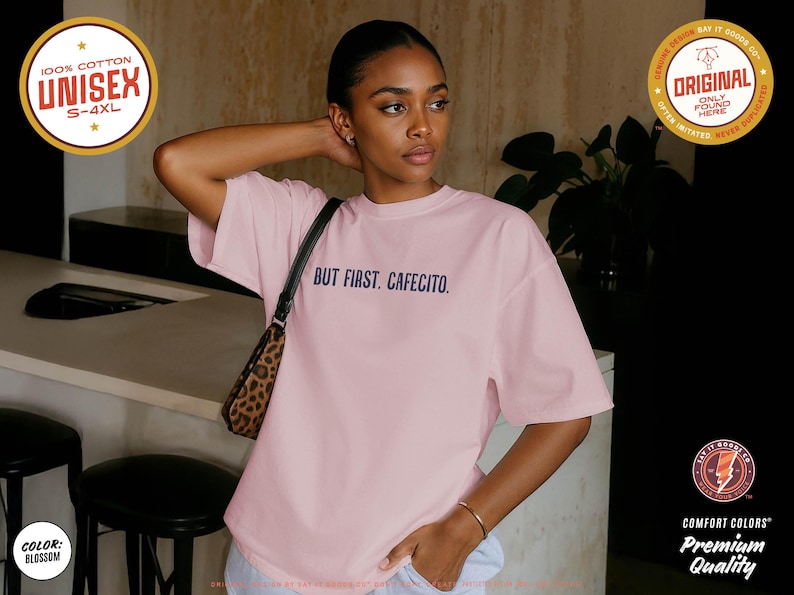 K&ouml;nnte beinhalten: Hellrosa Unisex-T-Shirt mit dem marineblauen Schriftzug "BUT FIRST. CAFECITO.". Das Shirt besteht aus 100 % Baumwolle und ist in den Gr&ouml;&szlig;en S-4XL erh&auml;ltlich. Es ist ein Premium-Qualit&auml;tsprodukt von Comfort Colors.