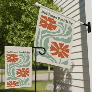Może przedstawiać: Dwie dekoracyjne flagi z napisem "Pollinate Positivity" i kwiatowym wzorem w kolorze pomarańczowym i zielonym. Jedna flaga jest zamontowana na słupie, druga na czarnym metalowym stojaku. Flagi są przeznaczone do użytku na zewnątrz.