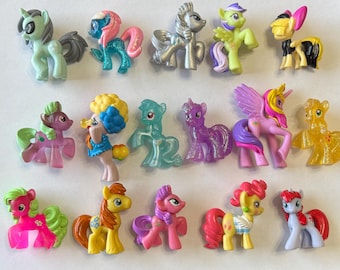 Puede incluir: Una colección de 16 pequeñas figuras de pony de plástico en varios colores y diseños. Los ponis están todos de pie sobre una superficie blanca.