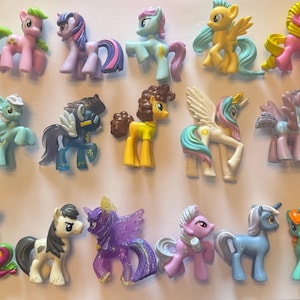 May include: A collection of fifteen small plastic pony figurines in various colours including pink, purple, blue, yellow, white, and green. The figures feature diverse manes, tails, and wing designs, with some characters displaying unique cutie marks on their flanks.