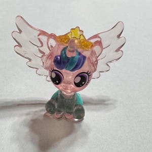 May include: A translucent pink and blue figurine of a winged unicorn with a gold crown. The toy has large, expressive eyes and is posed in a seated position. The wings are a lighter pink and the unicorn has a blue and purple mane.