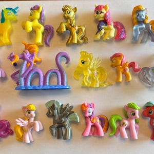 May include: A selection of colourful toy ponies in different poses. The ponies are in shades of yellow, pink, purple, and blue, with detailed features. Some have wings, and one is a sea serpent. The toys are made of plastic.
