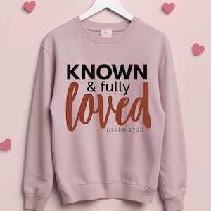Peut inclure: Un sweat-shirt rose clair avec l'inscription "KNOWN & fully loved" en noir et marron. Le texte "psalm 139:1" est en dessous de "loved". Le sweat-shirt est suspendu à un cintre blanc, avec des cœurs roses en arrière-plan.