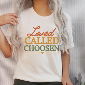 Op de afbeelding: Wit T-shirt met de woorden "Loved CALLED CHOOSEN" in oranje, geel en groen. Een klein hartje staat onder het woord "CHOOSEN". Het shirt heeft een ronde hals.